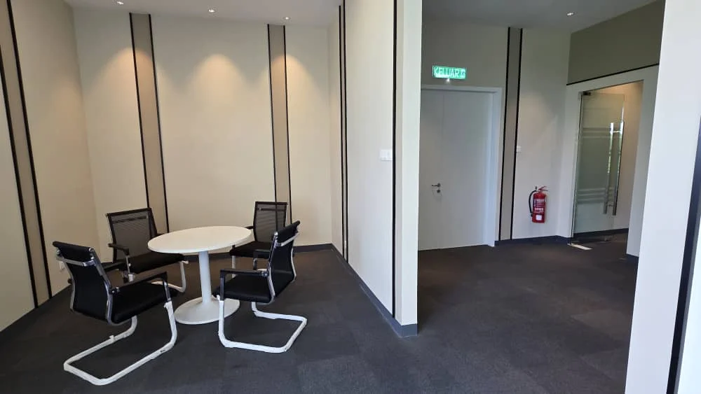 Room features: grey carpet flooring, white painted walls, recessed downlights, round white dining table, 4 black mesh chairs, glass partition wall, fire extinguisher mounted on wall, door with silver handle; style: modern.